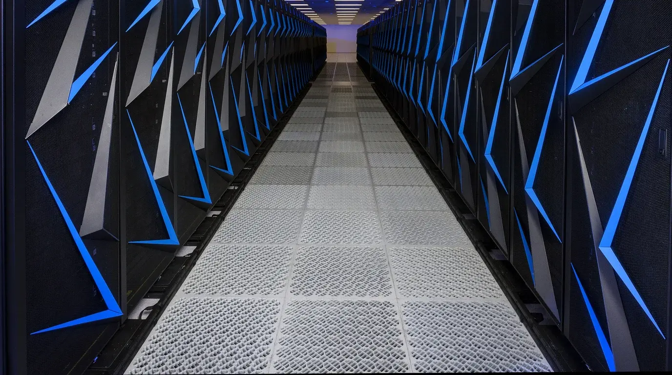 A long aisle between rows of black supercomputer cabinets marked with blue chevron light strips, a perforated floor running to the far wall.