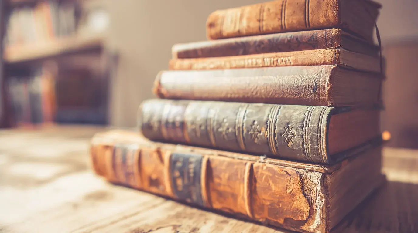 A stack of worn leather-bound books on a wooden table, a bookcase softly out of focus behind.