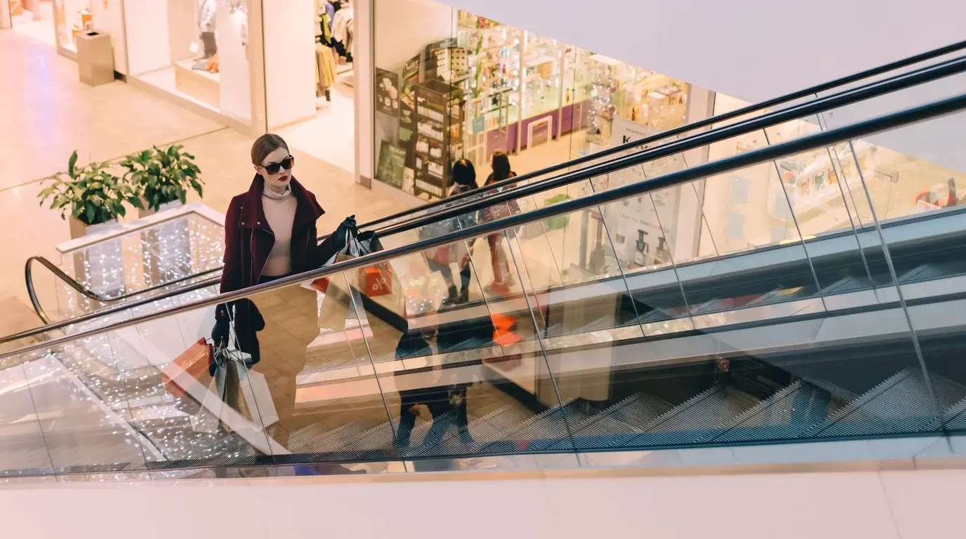 A woman in sunglasses carrying shopping bags rides an escalator in a bright shopping mall decorated with string lights.