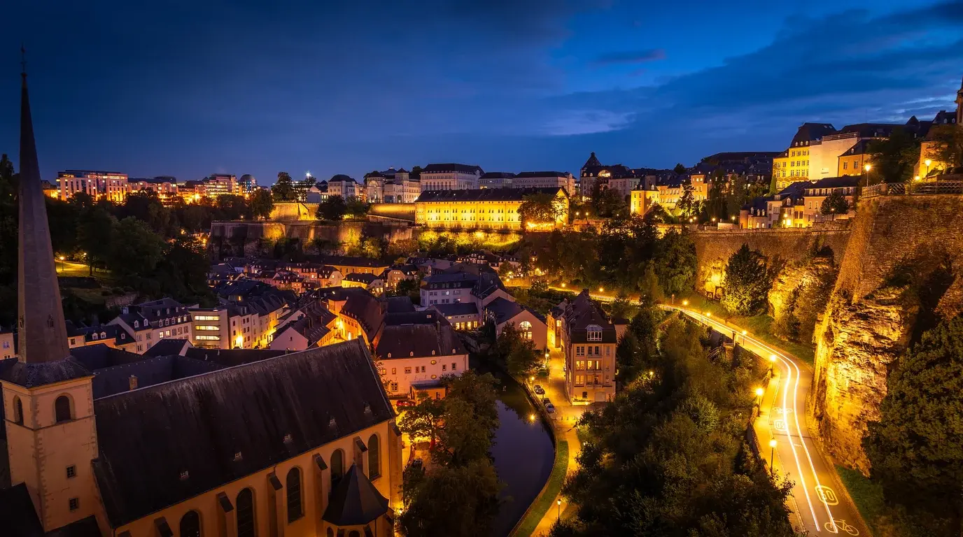 Luxembourg City's Grund quarter at dusk, a church spire and lit streets winding below the old-town cliffs.