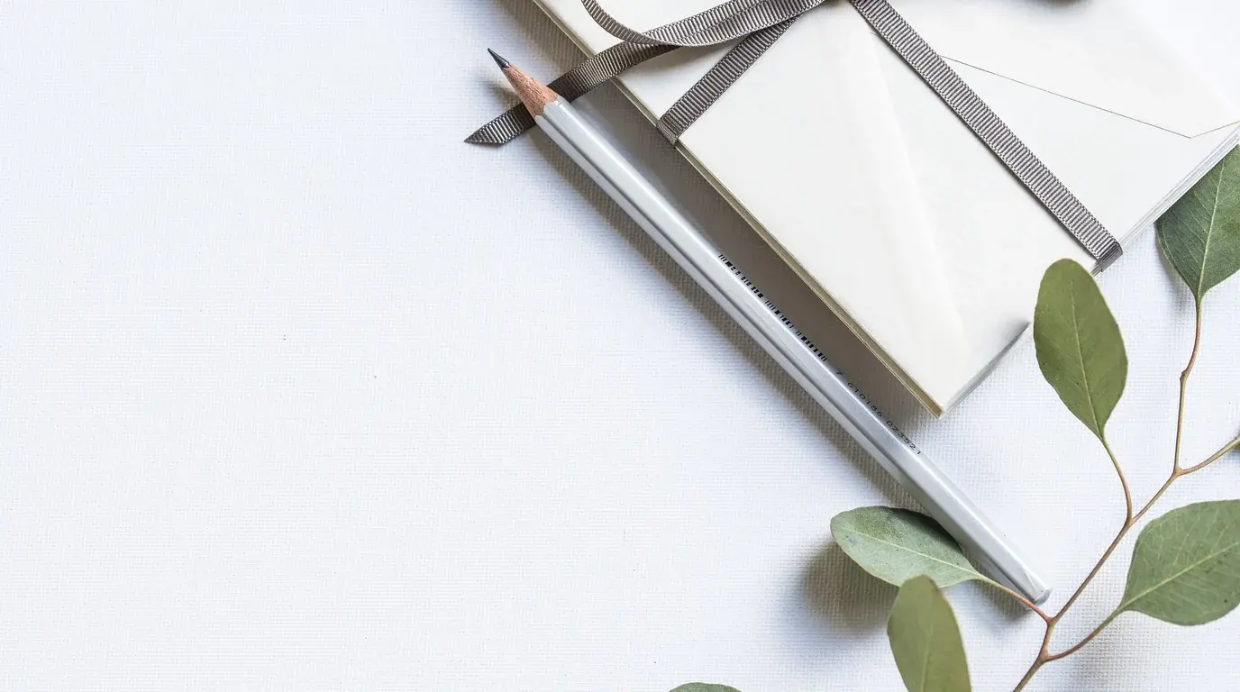 White envelopes tied with a grey ribbon beside a silver pencil and a eucalyptus sprig on white paper.