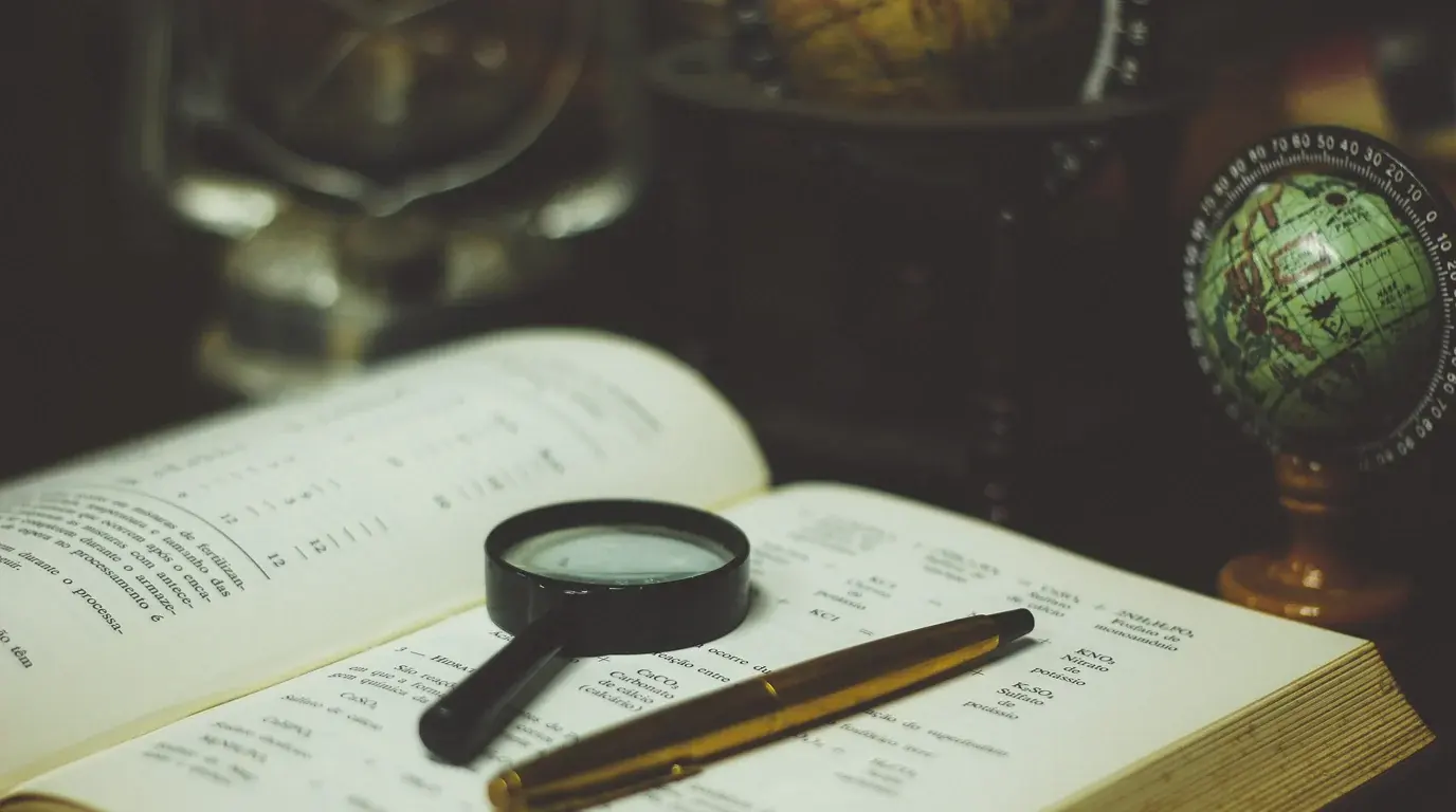 A magnifying glass and a gold pen resting on an open book, a small globe in the background.