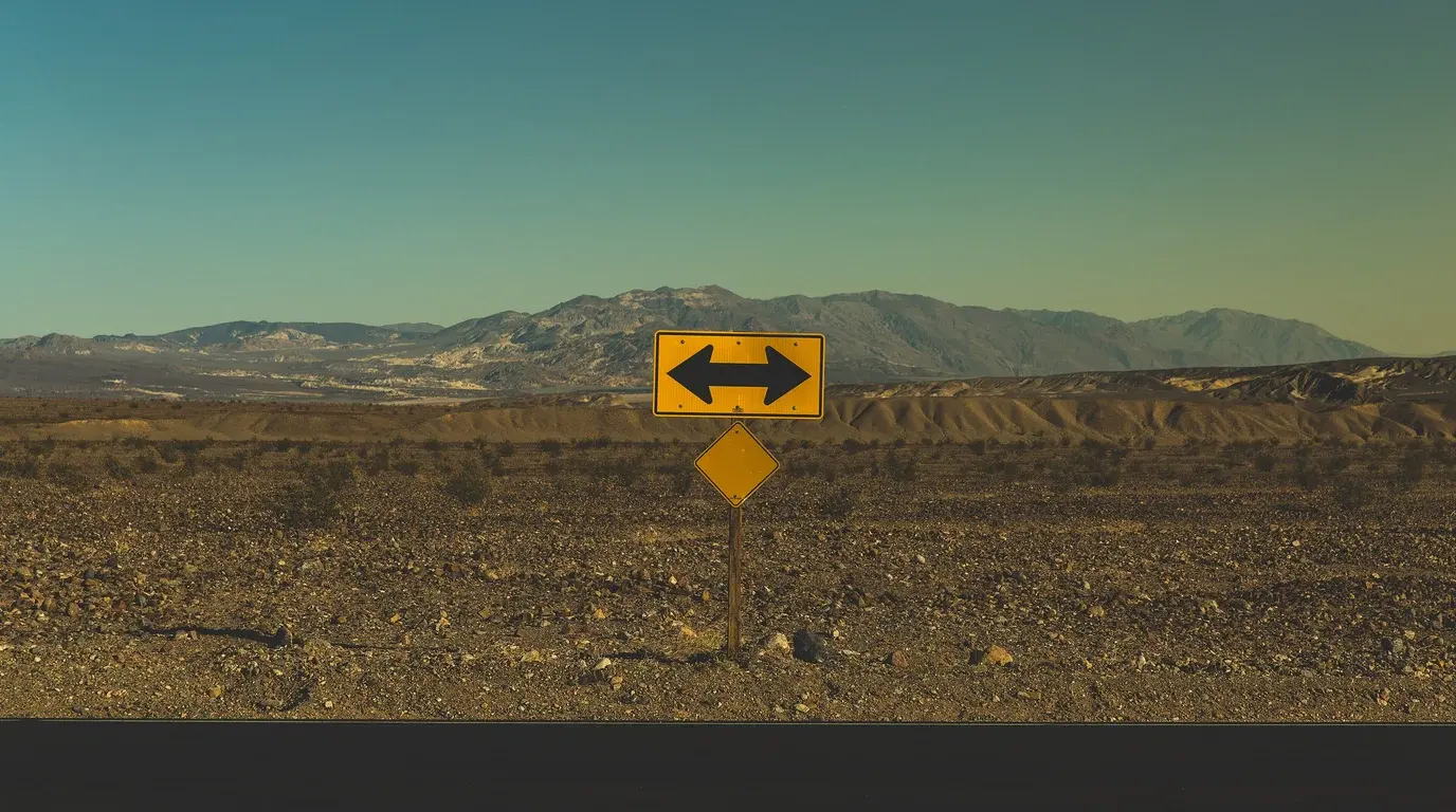 A yellow two-way arrow road sign on a post in a stony desert, mountains under a clear sky behind.
