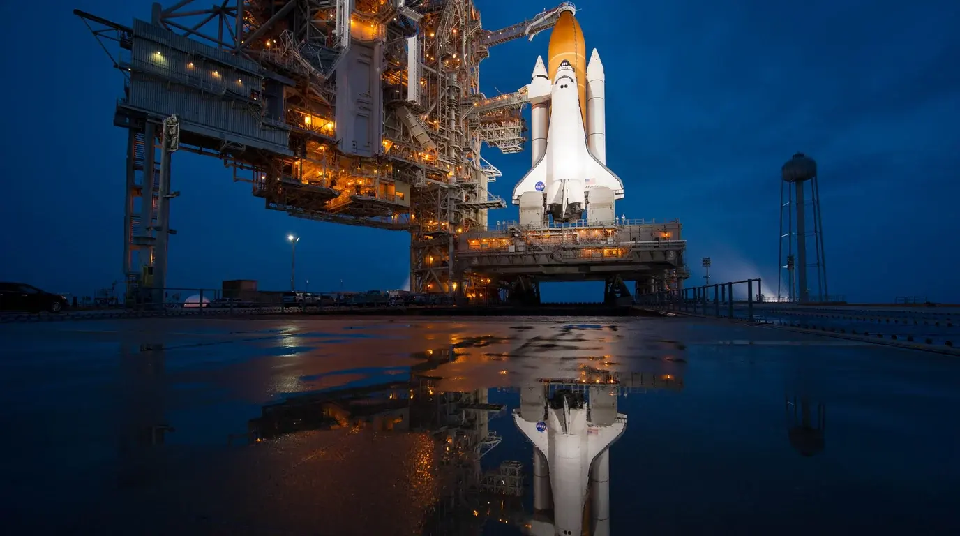 A space shuttle lit on its launch pad at dusk, tower and orbiter reflected in rainwater.