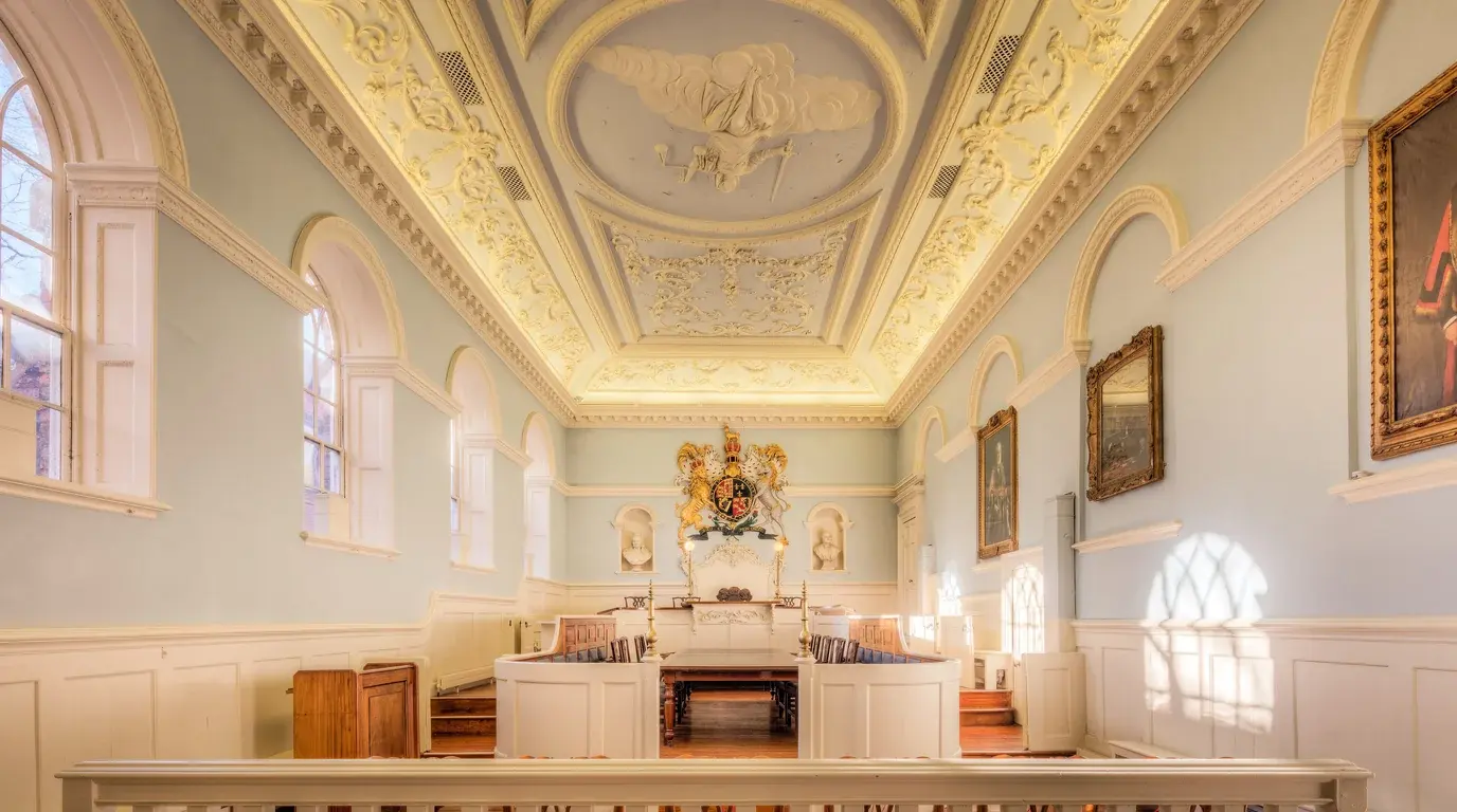 An ornate historic courtroom with a stuccoed ceiling, a royal coat of arms above the bench and tall arched windows.