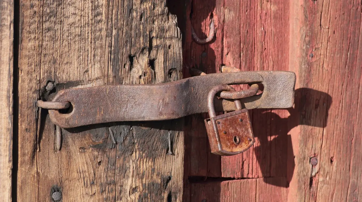 A rusted padlock closing an iron hasp on a weathered, red-painted wooden door.