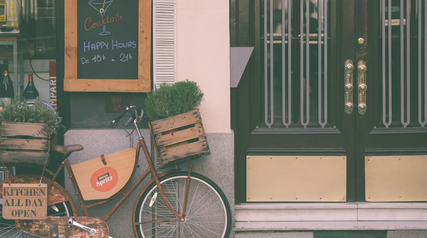 A small bar's street front with a chalkboard menu, bottles in the window and a bicycle carrying herb crates.