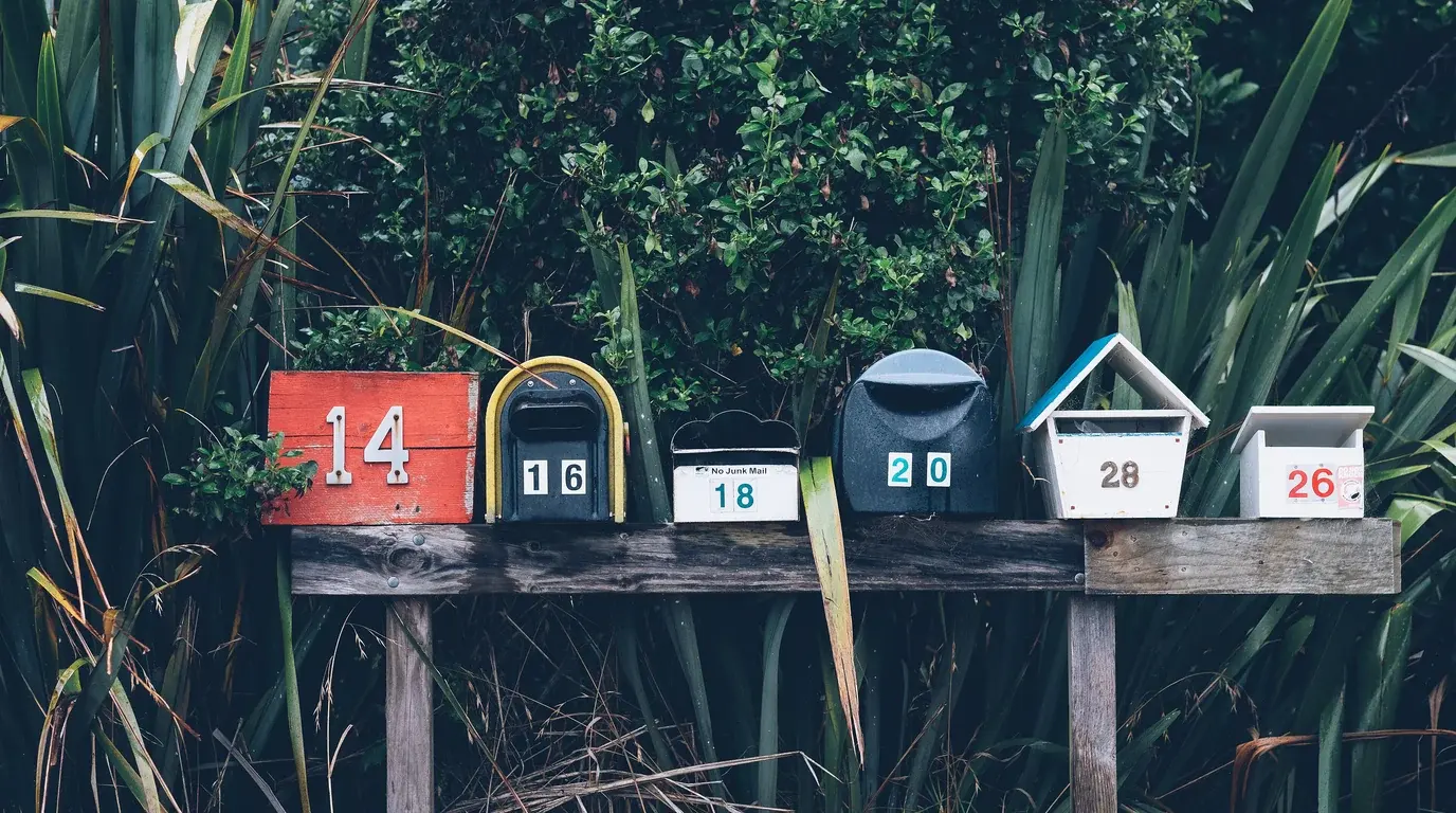 A row of six differently shaped, numbered mailboxes on a wooden bench in front of dense green foliage.