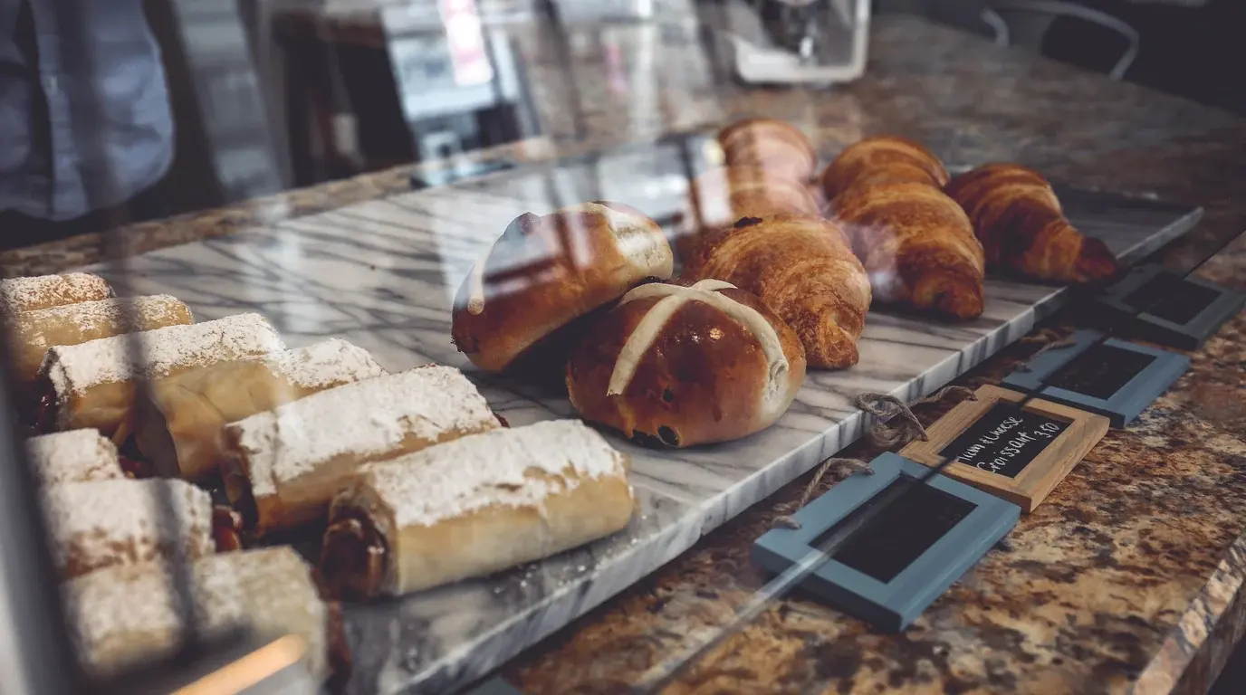 A bakery counter seen through glass, croissants, hot cross buns and sugared rolls on a marble tray.