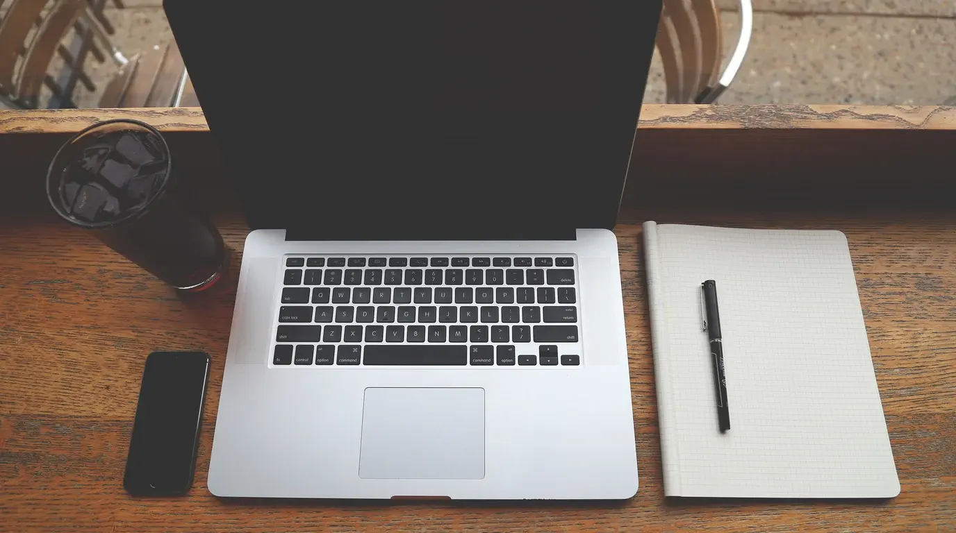 Top-down view of a laptop on a wooden desk between a smartphone, an iced drink and a notebook with a pen.