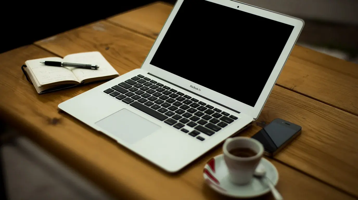 A laptop on a wooden desk beside an open notebook and pen, a smartphone and an espresso cup.