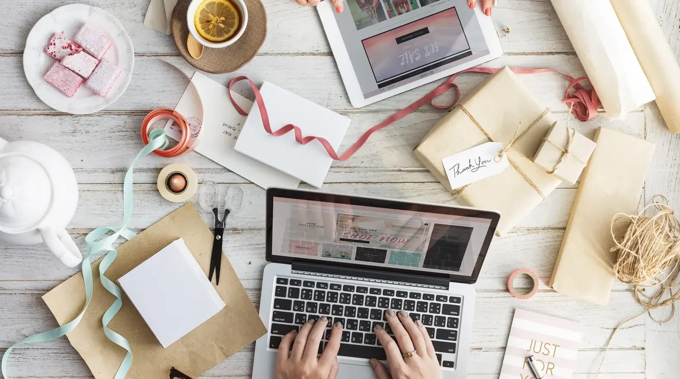 Top-down view of hands typing on a laptop among wrapped parcels, ribbons and a cup of tea on a white wooden table.