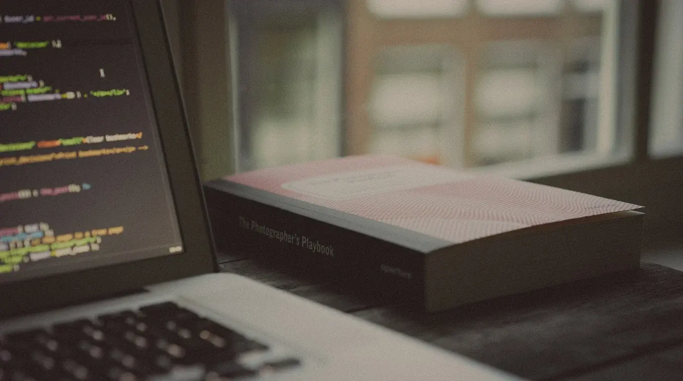 A laptop showing lines of code beside a closed book on a desk by a bright window.