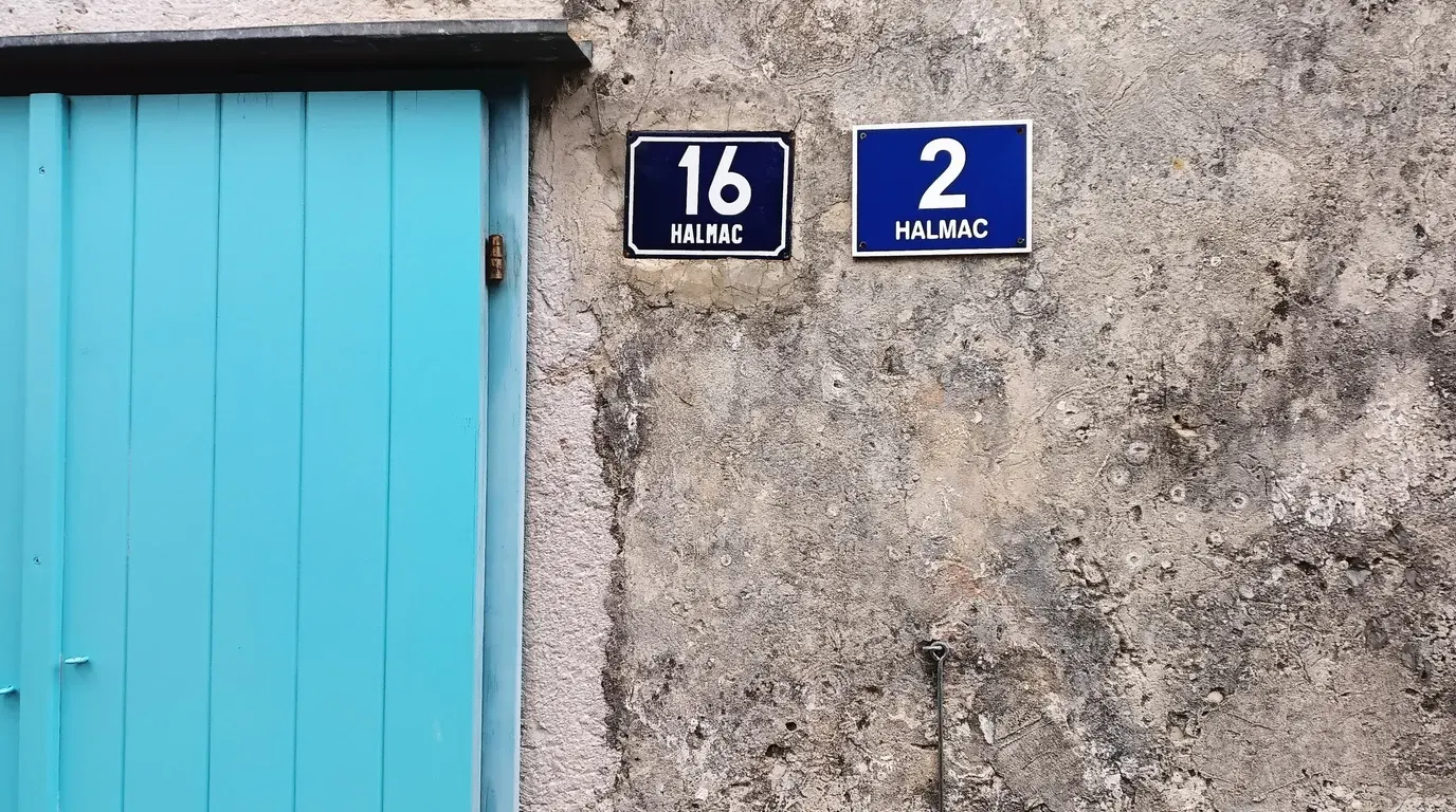 A turquoise wooden door beside two blue enamel house-number plates, 16 and 2, on a rough stone wall.