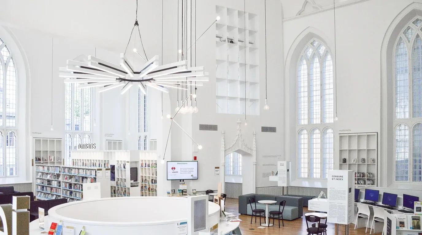 A bright white library hall with Gothic arched windows, a sculptural chandelier above a round information desk and open shelving.